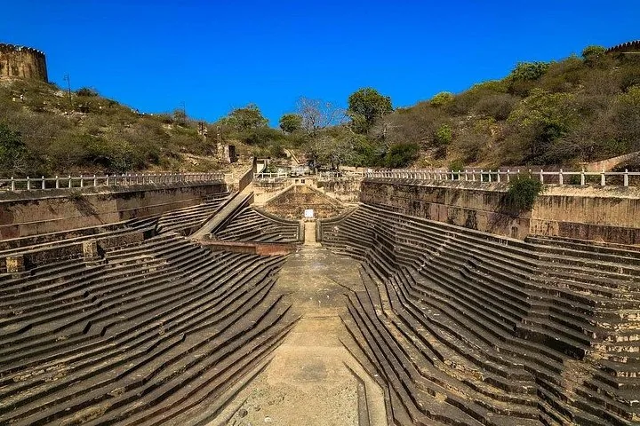 Un gran y antiguo pozo escalonado rodeado de colinas y murallas defensivas bajo un cielo azul.